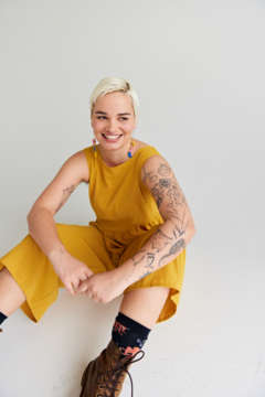 Becki wears a yellow jumpsuit and has tattoos. shes smiling