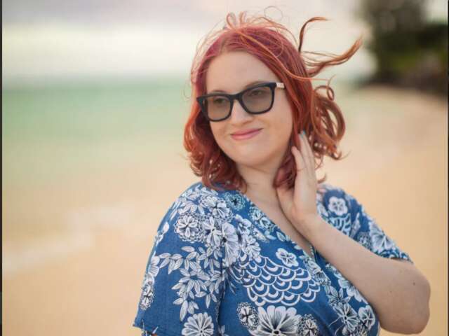 Natasha has dark red hair and fair-skin. She is standing at the beach wearing black sunglasses and a blue dress with white patterns.