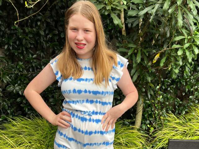 Molly has light hair and is wearing a white and blue striped dress. She is standing in front of a garden