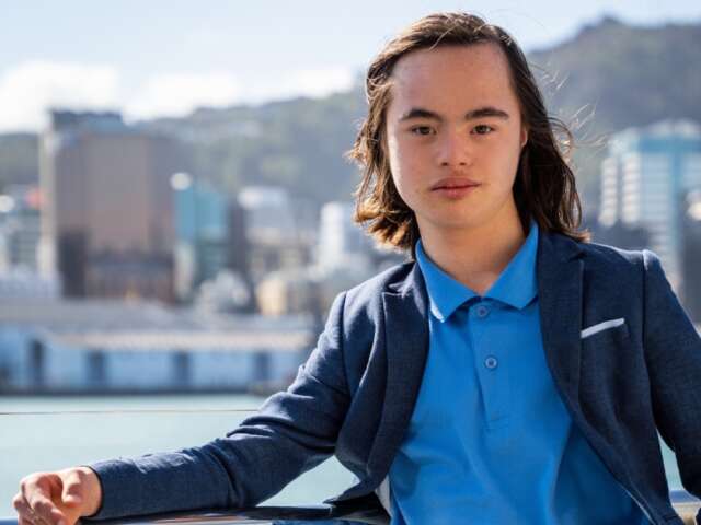Xavier is an asian male with long black hair. He is standing in a blue shirt and navy blazer. He is standing with the city in the background.