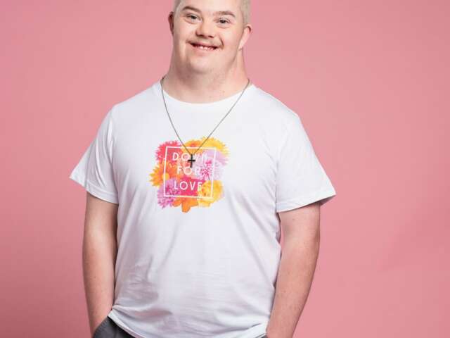Braydon is standing with a big smile wearing black pants and a white t-shirt with a colourful image on it. He is standing in front of a pink backdrop.