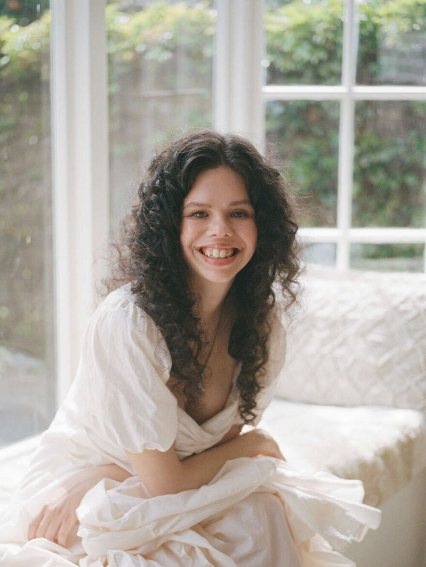 Mamie sits on her lounge day bed and wears a white dress with big curls