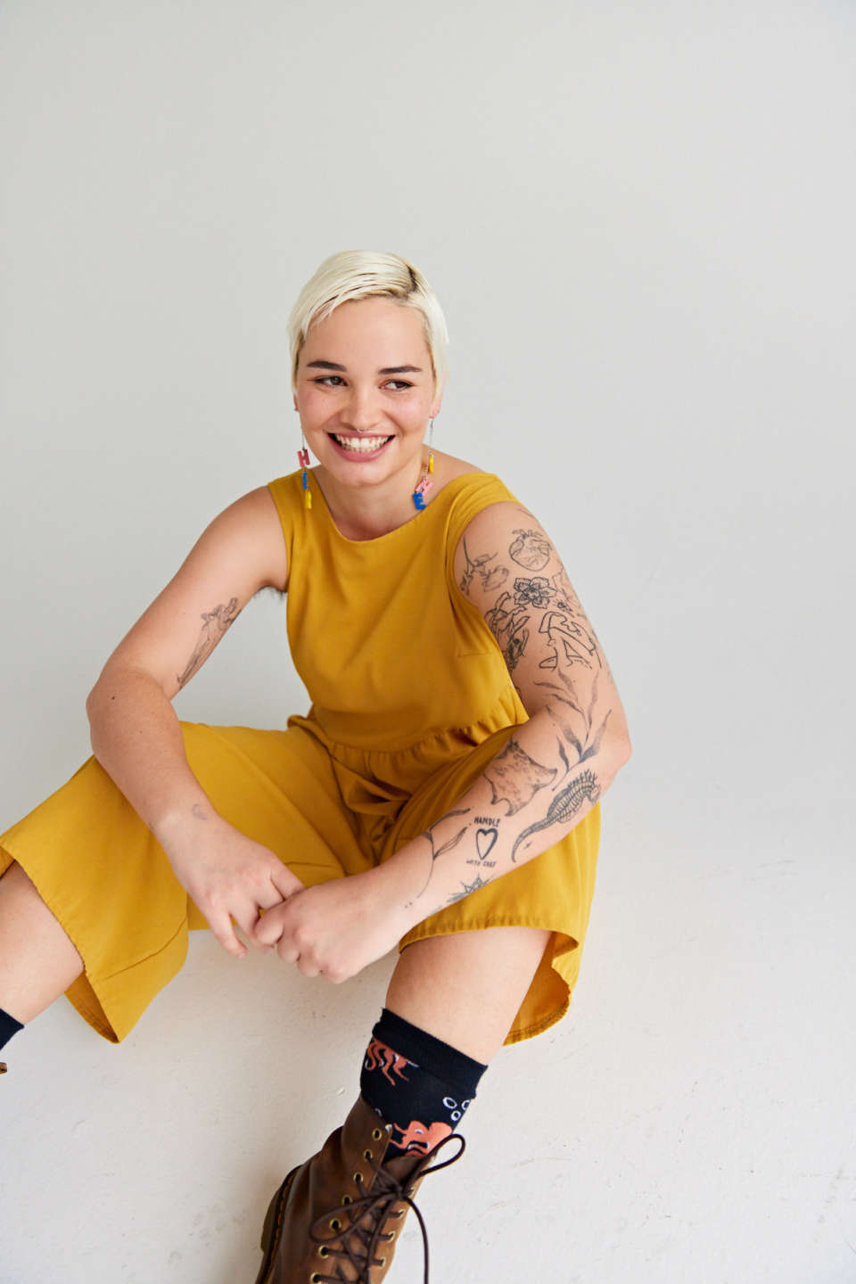Becki wears a yellow jumpsuit and has tattoos. shes smiling